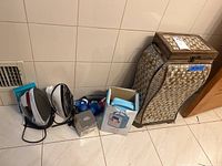 Group photo showing the painted wicker-style hamper, two electric irons, boxed blood glucose monitoring system, blood pressure monitor, and blue Songbird radio. Items arranged on tiled floor.