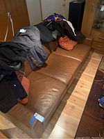Brown leather couch with some coats and clothing draped over, showing three seat cushions and armrests on both sides. Two pillows, one orange and one matching leather, visible on couch.