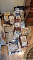 Photo showing assortment of packaged curtain panels, bed linens, and tablecloths arranged on floor beside a dresser.