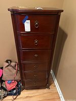 Front view of tall wooden lingerie chest with six drawers and metal ring pull handles showing the entire piece on a hardwood floor next to some bags.