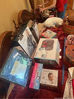 Photo showing various boxed and packaged items including a Homedics massager, cozy wrap, ankle and wrist weights, neck massager, and blanket stacked on a bed.