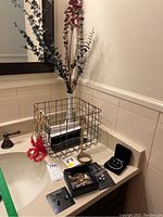 View of wire basket, white milk glass bud vase, assorted jewelry in black boxes and laid out on countertop with tags visible