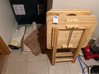 Five wooden snack trays stacked next to assorted throw rugs and mats on tile floor.