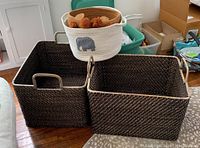 Two large rectangular dark woven baskets side by side on wooden floor with handles visible, showing woven pattern.