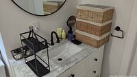 Full view of bathroom countertop showing woven baskets stacked, round mirror, magnifying mirror, soap dispenser, two-tier black metal shelf, and black wire basket with jars.