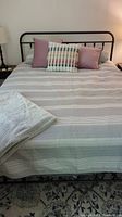 Full view of bed with striped grey and white sheets, coverlet, three toss pillows including light purple and plaid design, and fuzzy grey throw at the foot of the bed.