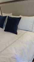 Bed showing white comforter and pillowcases with decorative chain-link embroidery and two navy blue square toss pillows.