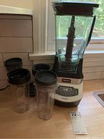 Blender base with large blending jar and four shaker cups with black lids on kitchen counter near window.