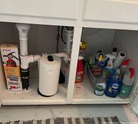 Under sink area with paper towel roll and assorted cleaning products in bottles and containers on white shelf, including Dawn dish soap and spray bottles.