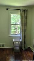 Full view of a single window with one grey curtain hung on a black metal rod and a rectangular hamper below the window.