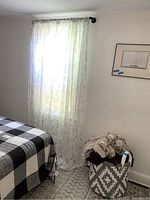 Full view of sheer curtains hanging on rod near window, part of bed and woven basket with blankets visible.