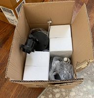 Top view of opened cardboard box with light fixture parts including black metal mounting plate and plastic wrapped components all secured with styrofoam inserts.