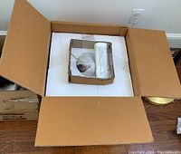 Box with protective styrofoam and cardboard inserts containing partially visible rolled instructions and glass component wrapped in plastic