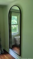Mirror in natural light reflecting window, curtain, and hardwood floor showing arched top and slim metal frame.