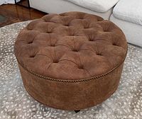 Round brown tufted ottoman on patterned rug in front of white couch showing top and front side.