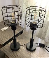 Pair of black metal desk lamps with cage-style shades on a wooden surface. Visible bulbs inside the cages.