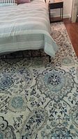 Partial view of the rug placed under a bed, showing medallion pattern in shades of grey and blue on a cream background.