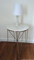 Full view of side table with marble top and metal base, with white table lamp on top, against a wall on wood floor.