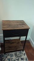 Side view of one brown wood finished side table with black metal frame and fabric drawer, showing height and structure against a light wall and carpeted floor.