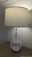 Full view of a single double gourd crackled glass table lamp with white cloth drum shade on wooden table, lit side angle.