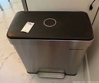 Full view of rectangular stainless steel trash can with black plastic lid and foot pedal placed on a marble floor next to a wall.
