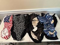 Multiple one-piece swimsuits laid out on a table showing various patterns including American flag, geometric black and white, striped, zebra print, and black solid.