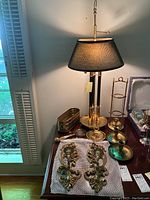 Brass lamp with black shade, brass candle holders, brass container, and magnifying lens displayed on a wooden table.