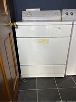 Front and side view of white Whirlpool dryer positioned next to a washing machine, showing front door with handle and appliance on a tile floor.
