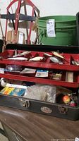 Photo showing vintage fishing tackle box open with fishing lures, hooks, weights, bobbers, and fishing line inside, next to a green bucket.