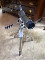 Full view of matte black Tasco spotting scope mounted on silver Rokinon tripod on beige carpet.