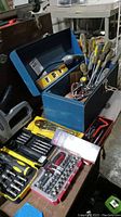 Blue metal tool box open with variety of hand tools inside, including screwdrivers, wrenches, and socket sets visible on table.
