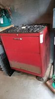 Red heavy metal toolbox with caster wheels and three drawers, with assorted metal tools visible on and inside the drawers.