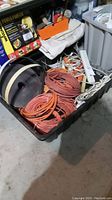 View of tote showing six large orange extension cords coiled inside along with white surge protector strips and miscellaneous cords, plus large heavy black roll of material.