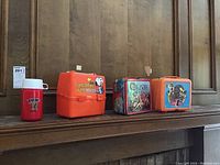 All lunch boxes and thermos arranged on mantle, showing Snoopy lunch boxes, Thundercats metal lunch box, Mr T plastic lunch box and thermos