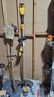 Dewalt yellow and black weed eater and charger standing upright against a wooden wall with other tools.