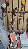 Photo showing two gas operated garden tools (weed eater and edger) hanging on a wall with a box of parts below.