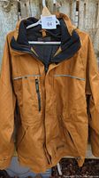 Front view of tan Dakota men's jacket hanging on a wooden hanger outdoors showing the front zipper and zippered chest pocket.