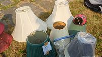 Multiple assorted lamp shades placed on grass, showing white fabric shades, green pleated shade, small red and black decorative shade, and wrapped shades.