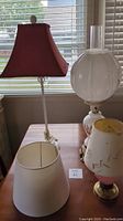 Tall table lamp with twisted clear base and red fabric shade, white opaque glass lamp base, small pink frosted glass lamp with brass base and floral shade, and white lamp shade on table near window.