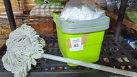 Photo of a new mop and bucket set. Green bucket with a wringer lid and mop with white cotton strands and a silver handle placed next to it.
