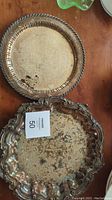 Photo of the top of the two silver platters stacked, showing decorative edge of the smaller round platter and ornate border of the larger platter with tarnish spots and discoloration