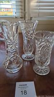 Four clear crystal vases arranged on a wooden surface near window light, showing various cut and etched designs.