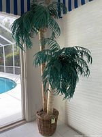 8-foot tall faux palm tree with multiple green fronds and textured trunk inside a woven basket, placed indoors next to a sliding glass door by a pool area.