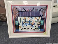 Framed artwork with white frame and glass, showing a village scene with stylized elements including two human figures, birds, trees, and architectural shapes. The frame sits on the floor against a wall with surroundings visible.