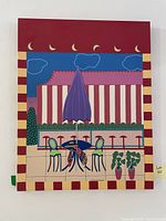 Full view of colorful painting showing outdoor café scene with umbrella, table, chairs, and decorative elements.