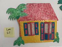 Metal wall art showing a tropical house with red roof, yellow walls, blue painted window and door frames with white shutters, flanked by green palm trees.