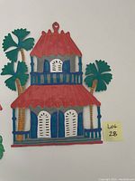 Photo of a metal wall hanging art shaped like a house with red roof and blue railings, flanked by two green palm trees.