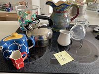 Photo showing assortment including decorative pitchers, creamers, stainless steel kettle, and clear container with fish design on stove top.