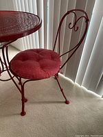 Single red metal chair with curved scrollback and red cushioned seat on light carpet beside vertical blinds.