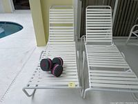 Two white chaise loungers side by side on a pool deck with a pair of pink and black hand weights resting on one lounger.